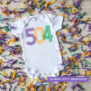 Puede incluir: Un body blanco para bebé con el número 504 impreso en verde, amarillo y morado. El body está rodeado de confeti morado, verde y amarillo. El texto "QUEEN CITY BESPOKE" está impreso debajo del body.