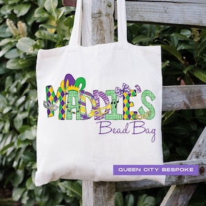 K&ouml;nnte beinhalten: Eine wei&szlig;e Canvas-Tragetasche mit dem Namen "Maddie's" in violetten und gr&uuml;nen gemusterten Buchstaben. Die Buchstaben sind mit Mardi Gras-Motiven verziert. Der Text "Bead Bag" ist unter dem Namen in Lila gedruckt. Der Text "Queen City Bespoke" ist unten rechts gedruckt.