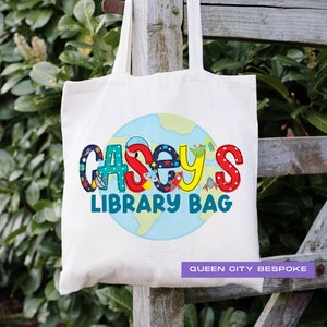 Może przedstawiać: Biała torba z płótna z kolorową grafiką globusa i tekstem "Casey's Library Bag" w zabawnym kroju pisma. Torba ma również napis "Queen City Bespoke" w prawym dolnym rogu.
