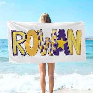 以下が含まれることがあります： 白いビーチタオルに、大きなカラフルな文字で「ROWAN」と書かれています。文字は紫で縁取られ、黄色、紫、白のパターンで塗りつぶされています。単語の真ん中には黄色の星があります。