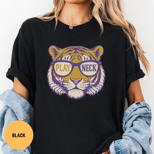 May include: Black t-shirt featuring a tiger face graphic wearing sunglasses. The sunglasses have the words "PLAY" and "NECK" printed on them. The tiger graphic is in shades of purple, gold, and cream.