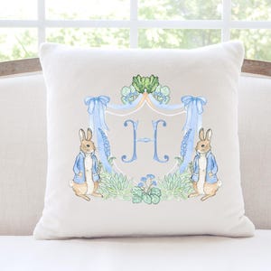 May include: A square, off-white cushion with a watercolour design. The design features two rabbits in blue jackets either side of a light blue initial "H" within a decorative crest. Green foliage and blue ribbons complete the design.