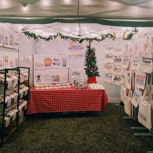 May include: A Christmas market stall with a variety of handmade items for sale, including tote bags with funny sayings, tea towels, and small boxes. The stall is decorated with a small Christmas tree and string lights. The sign above the stall reads "Queen City Bespoke".