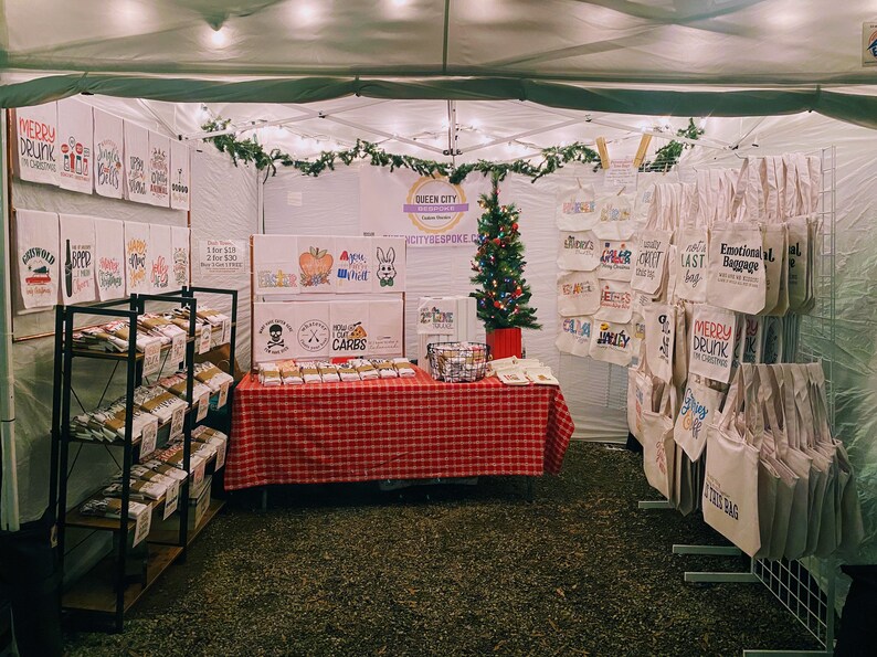 May include: A Christmas market stall with a variety of handmade items for sale, including tote bags with funny sayings, tea towels, and small boxes. The stall is decorated with a small Christmas tree and string lights. The sign above the stall reads "Queen City Bespoke".