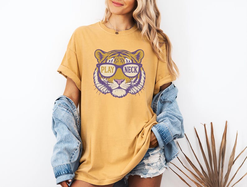May include: Mustard yellow t-shirt featuring a tiger face graphic wearing sunglasses. The tiger graphic is purple and gold, with the words "PLAY NECK" printed on the design. The shirt has short sleeves and a relaxed fit.