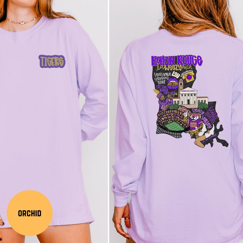 May include: Orchid-colored long-sleeve shirt with the word "TIGERS" in purple on the front. The back of the shirt features a graphic of Louisiana with the words "Baton Rouge" and other text in purple and gold.