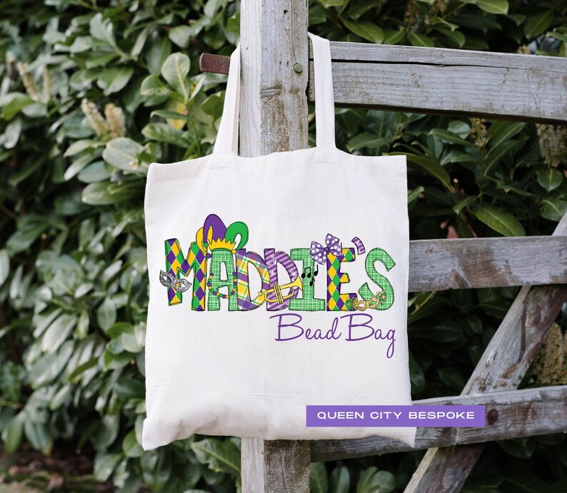 K&ouml;nnte beinhalten: Eine wei&szlig;e Canvas-Tragetasche mit dem Namen "Maddie's" in violetten und gr&uuml;nen gemusterten Buchstaben. Die Buchstaben sind mit Mardi Gras-Motiven verziert. Der Text "Bead Bag" ist unter dem Namen in Lila gedruckt. Der Text "Queen City Bespoke" ist unten rechts gedruckt.