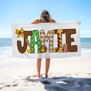 May include: A white beach towel with the name "JAMIE" spelled out in colorful letters. The letters are decorated with baseball and softball imagery, including bats, balls, and a popcorn bucket.