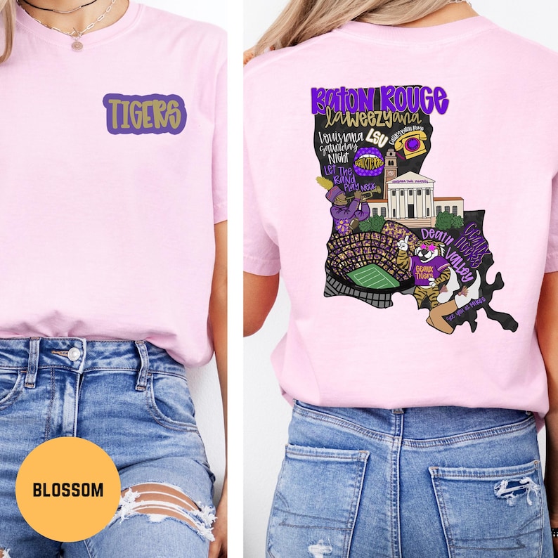 May include: Light pink t-shirt with the word "TIGERS" in purple and gold on the front. The back features a purple and black graphic of Louisiana with "Baton Rouge, Louisiana" and other text.