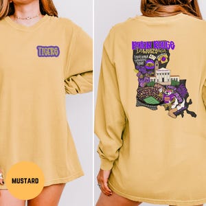 May include: Mustard-colored long-sleeve shirt with "TIGERS" in purple on the front. The back features a purple and white graphic of Louisiana with "Baton Rouge" and other text. The shirt is a comfortable, casual style.