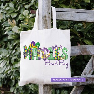 K&ouml;nnte beinhalten: Eine wei&szlig;e Canvas-Tragetasche mit dem Text "Maddie's Bead Bag" in Lila, Gr&uuml;n und Gold. Der Text ist mit Mardi Gras-Grafiken verziert, darunter eine Narrenkappe, Noten und Masken. Die Tasche tr&auml;gt auch den Text "Queen City Bespoke" in Schwarz.