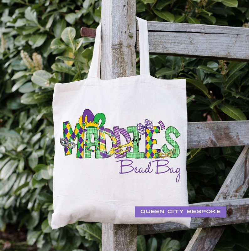K&ouml;nnte beinhalten: Eine wei&szlig;e Canvas-Tragetasche mit dem Text "Maddie's Bead Bag" in Lila, Gr&uuml;n und Gold. Der Text ist mit Mardi Gras-Grafiken verziert, darunter eine Narrenkappe, Noten und Masken. Die Tasche tr&auml;gt auch den Text "Queen City Bespoke" in Schwarz.