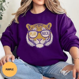 May include: A purple sweatshirt featuring a tiger face wearing sunglasses with the words "PLAY NECK" in the lenses. The tiger design is in shades of gold, tan, and white. The sweatshirt is a solid purple color.