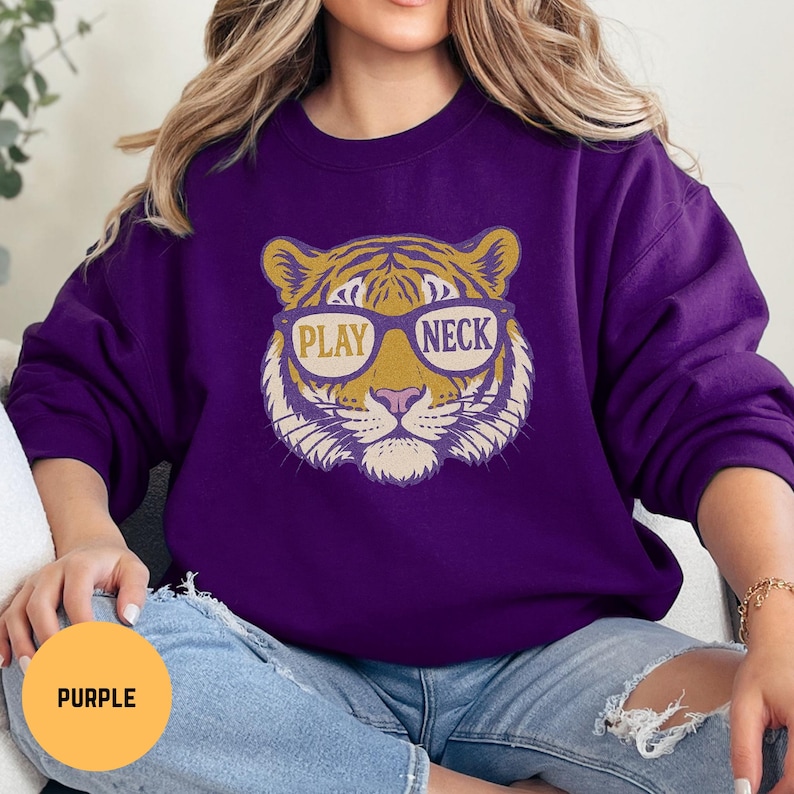 May include: A purple sweatshirt featuring a tiger face wearing sunglasses with the words "PLAY NECK" in the lenses. The tiger design is in shades of gold, tan, and white. The sweatshirt is a solid purple color.