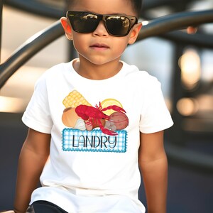 May include: A white t-shirt with a blue and white gingham design and a graphic of a crawfish, corn, lemon, and other ingredients. The text "Landry" is printed on the shirt.