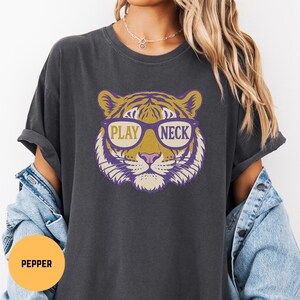 May include: Dark gray t-shirt featuring a tiger face wearing glasses with the words "PLAY NECK" in a gold and purple design. The tiger's face is detailed with yellow, purple, and white accents. The shirt has short sleeves.