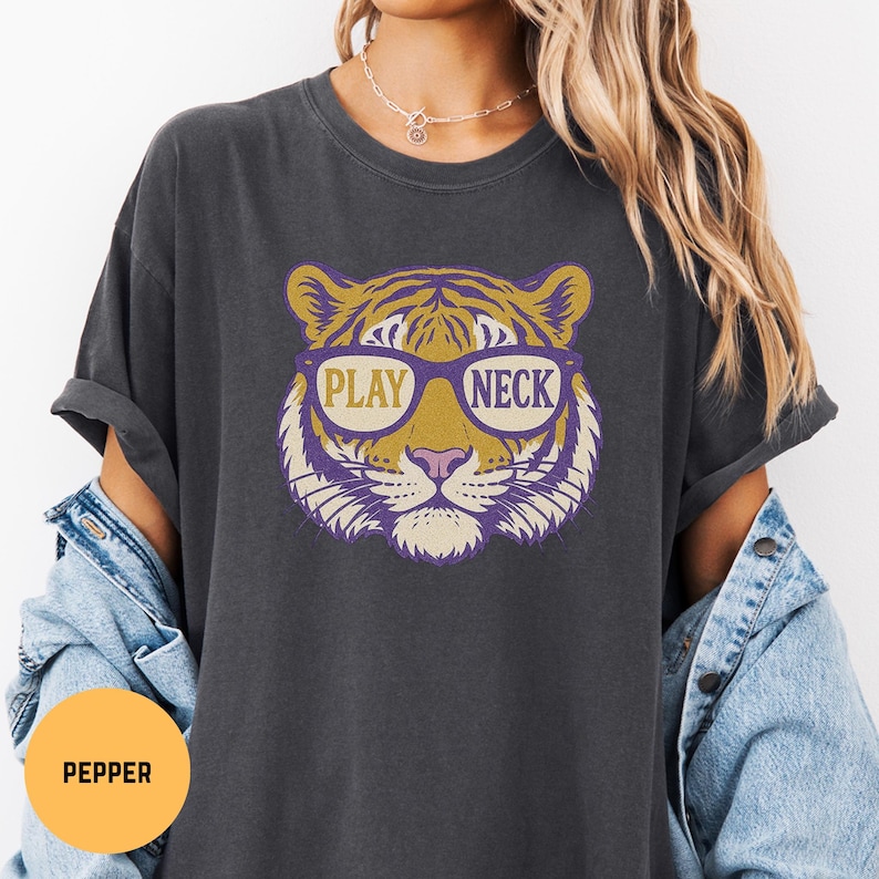 May include: Dark gray t-shirt featuring a tiger face wearing glasses with the words "PLAY NECK" in a gold and purple design. The tiger's face is detailed with yellow, purple, and white accents. The shirt has short sleeves.
