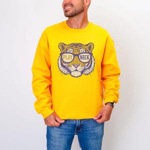 May include: A yellow sweatshirt featuring a tiger face graphic wearing glasses with the words "PLAY NECK". The graphic is in shades of purple, tan, and white. The sweatshirt is long-sleeved and is being worn with blue jeans.