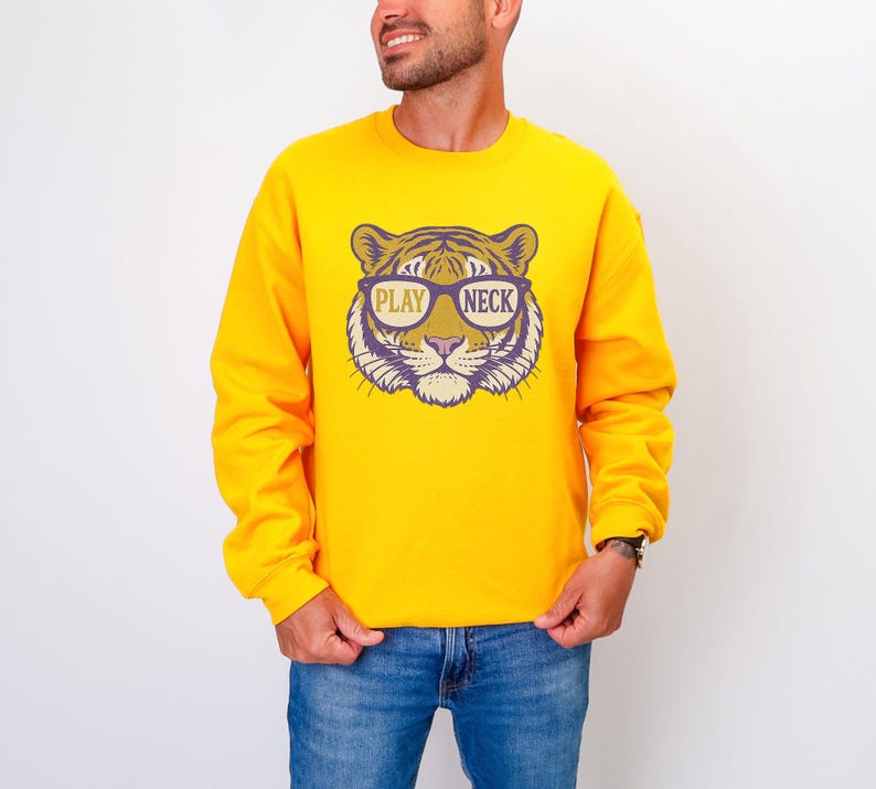 May include: A yellow sweatshirt featuring a tiger face graphic wearing glasses with the words "PLAY NECK". The graphic is in shades of purple, tan, and white. The sweatshirt is long-sleeved and is being worn with blue jeans.