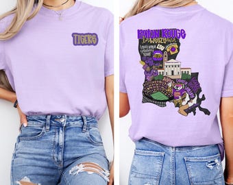 May include: A light purple t-shirt with the word "TIGERS" in a purple and gold design on the front. The back features a graphic of Louisiana with text and images related to Baton Rouge and LSU.