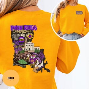 May include: Gold sweatshirt with a graphic of the Louisiana state outline, featuring text and illustrations related to Baton Rouge and LSU. The sweatshirt has the word "Tigers" on the upper right chest.