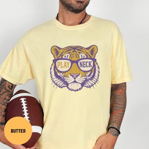 May include: Butter-colored t-shirt featuring a tiger head wearing glasses with the words "PLAY NECK" in a purple and gold design. The shirt is paired with a brown football.