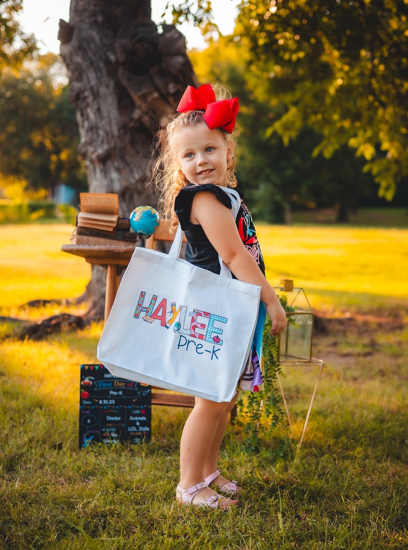 May include: A young girl with a red bow in her hair is holding a white tote bag with the name "HAYLEE" and "Pre-K" written on it in colorful letters. The bag is decorated with school-themed images.