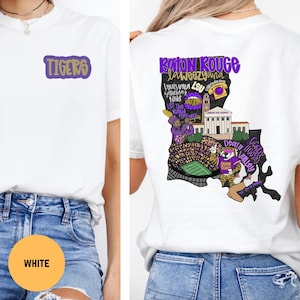 May include: White t-shirt with a purple and gold "TIGERS" logo on the front. The back features a purple and gold design with text including "Baton Rouge", "Louisiana", and "Death Valley".