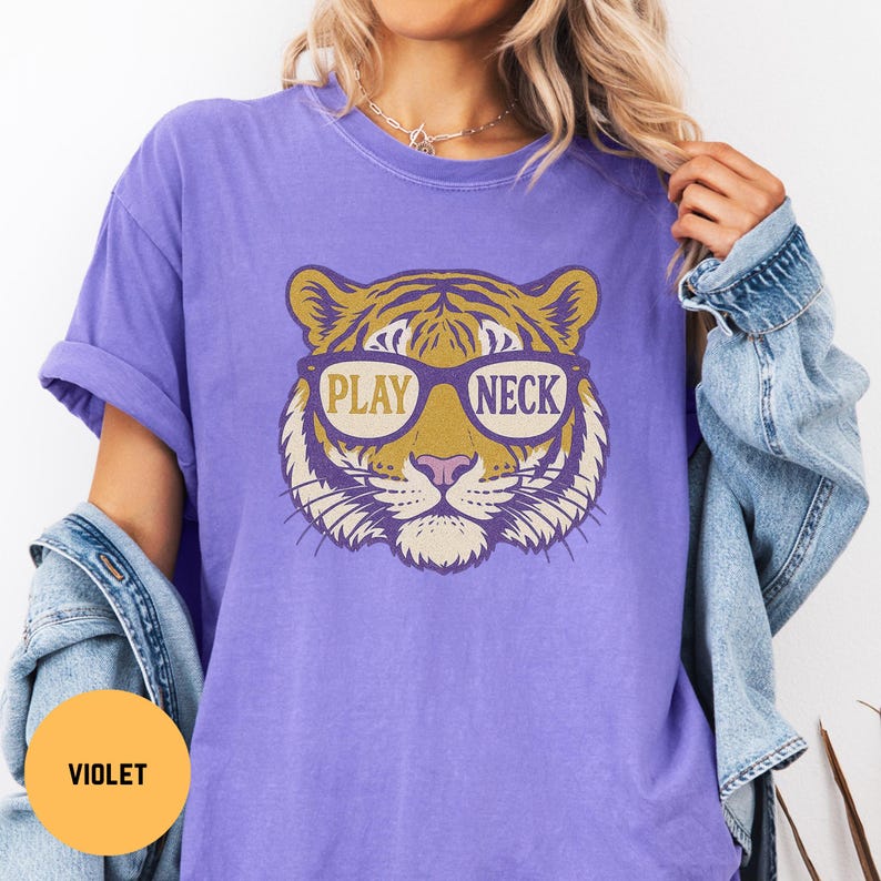 May include: A violet t-shirt features a tiger face graphic wearing glasses with the words "PLAY NECK". The tiger has gold and purple stripes, a pink nose, and is set against a violet background. The shirt has rolled-up sleeves.
