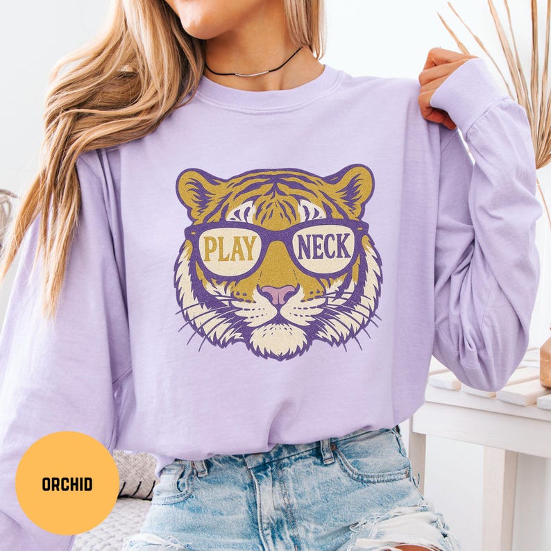 May include: A light purple long-sleeve shirt featuring a tiger face wearing glasses. The glasses have the words "PLAY NECK" printed on them. The tiger face is yellow, purple, and white.