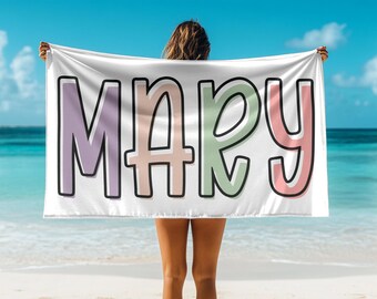 May include: A white beach towel with the name "MARY" written in colorful, outlined letters. The letters are in shades of pink, purple, green, and peach.