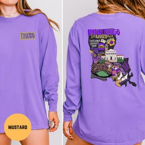 May include: A long-sleeved purple shirt with the word "TIGERS" in gold lettering on the front. The back of the shirt features a graphic design with the words "Baton Rouge laweezyana" and other text and images in purple, gold, and black.