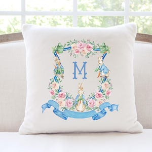 May include: A square white cushion with a floral wreath design, including pink roses, blue ribbons, and rabbit illustrations. A large blue letter "M" is at the centre. The cushion is on a white sofa.
