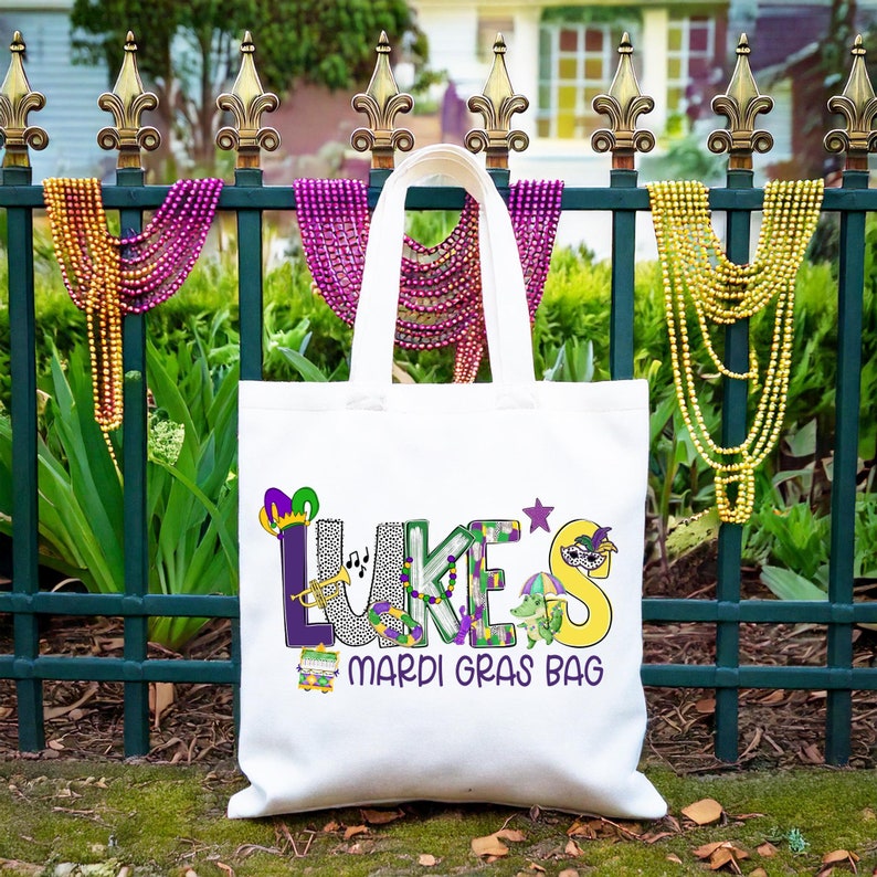 May include: White tote bag with the text "LUKE'S Mardi Gras Bag" in colorful letters with Mardi Gras themed decorations. The bag is hanging on a green metal fence with purple, gold, and green beads.