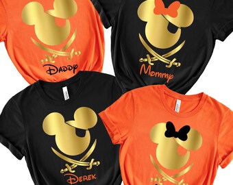 May include: Four t-shirts in black and orange feature a gold Mickey Mouse design with crossed swords. The shirts are labeled "Daddy", "Mommy", "Derek", and "Molly". The shirts are knotted at the bottom.