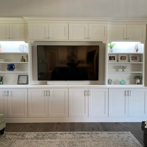 May include: A white entertainment center with a large flat screen TV, shelves, and cabinets. The unit has a modern design with sleek lines and simple hardware.