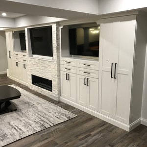 May include: A white entertainment center with a fireplace and three televisions. The entertainment center has multiple cabinets and drawers with black handles. The room has a gray rug and a black coffee table.