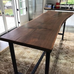 May include: A dark brown live edge walnut wood countertop with a black metal base. The countertop has a natural, organic shape and is supported by a simple, industrial-style base.