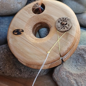 Może przedstawiać: Drewniane narzędzie do wiązania much z otworem w środku i małym guzikiem z boku. Narzędzie jest wykonane z jasnobrązowego drewna i ma przymocowaną białą sznurówkę.