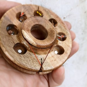Puede incluir: Una herramienta de atado de moscas de madera con seis ranuras para sujetar las moscas. La herramienta tiene un hilo rojo envuelto alrededor y una mosca está unida al hilo.