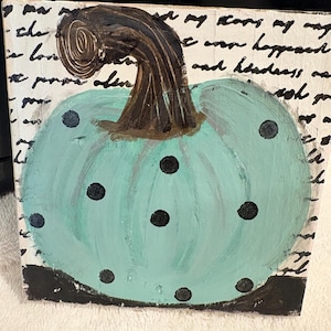 May include: A square canvas painting featuring a light teal pumpkin with black polka dots and a dark brown stem. The background consists of white painted surface with black cursive handwriting script and a solid black horizontal base line.