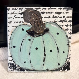 May include: A square canvas painting featuring a light mint green pumpkin with black polka dots and a dark brown stem. The background consists of white painted surface with black cursive script text and black textured accents at the bottom corners.