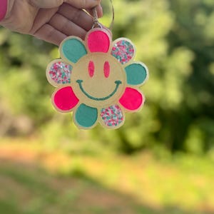 May include: A wooden flower ornament with a smiley face in the center. The flower is painted pink and teal with glitter accents.