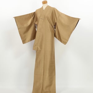 May include: A solid tan kimono featuring wide, extended sleeves and a wrap-front design. The garment is displayed on a mannequin, showcasing a floor-length silhouette with a clean, unpatterned surface and a structured collar that folds at the neck.