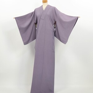 May include: A solid light purple kimono featuring wide, flowing sleeves and a traditional wrap-front design. The garment is displayed on a mannequin, showcasing its full-length silhouette, clean lines, and smooth, matte fabric texture throughout.