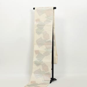 Può includere: Un tessuto color crema con un motivo ripetuto di forme astratte nei toni rosa, verdi e grigi tenui. Il tessuto è esposto su un supporto nero, che ne mette in risalto la lunghezza e il design. Sembra essere un oggetto decorativo, forse una sciarpa o uno scialle.