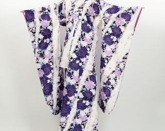 May include: A white kimono with a floral pattern of purple and lavender roses. The kimono has long, wide sleeves and a wrap-around design. The floral pattern is set against white stripes.