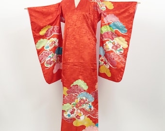 May include: A bright red kimono with a textured pattern and colourful cloud and floral designs. The kimono is displayed on a mannequin, showcasing its long sleeves and traditional V-neck. The decorative elements include shades of yellow, pink, green, and blue.