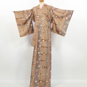 May include: A tan kimono with a striped and floral pattern. The kimono has wide sleeves and a V-neck. The fabric features vertical stripes, floral motifs, and geometric designs in shades of brown, gray, and red. The kimono is displayed on a mannequin.