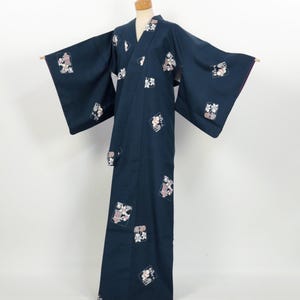 May include: A dark navy blue kimono with square floral patterns in pink, white, and grey. The kimono has wide sleeves and a wrap-around design, displayed on a mannequin. The edges of the sleeves are trimmed with a purple fabric.